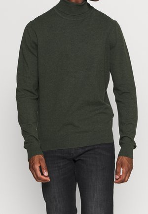 Homme portant un pull à col roulé vert foncé et un pantalon noir, debout devant un fond clair uni.