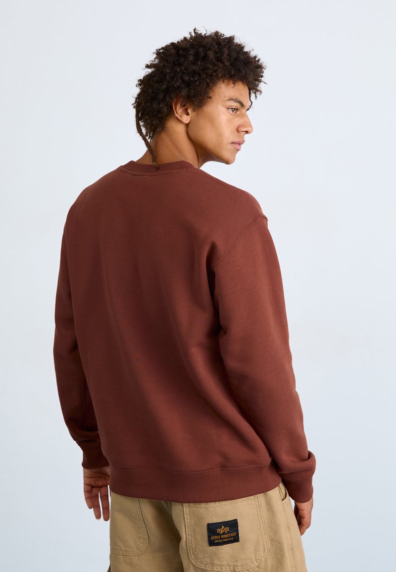 Brauner Sweatshirt mit Rundhalsausschnitt und gerippten Bündchen, von hinten gezeigt, kombiniert mit khakifarbenen Hosen mit einem Logo-Patch.