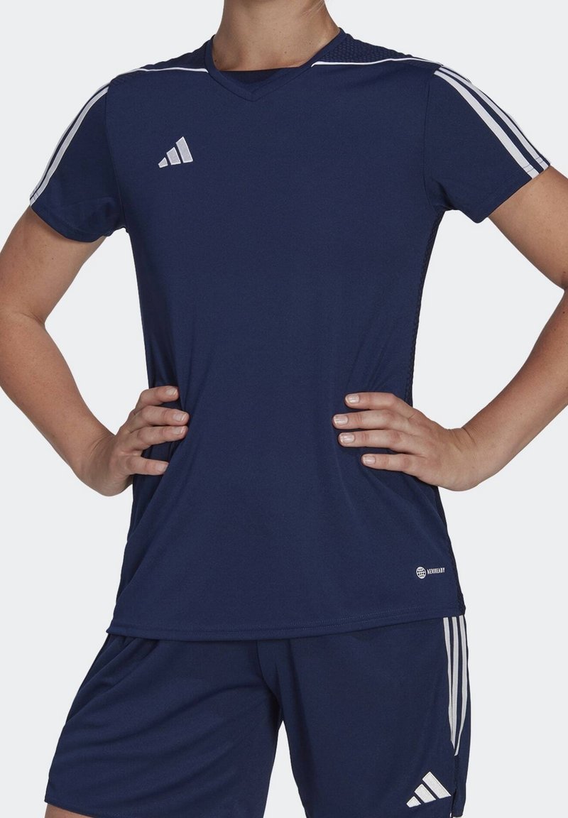 Laivastonsininen urheilupaita V-aukolla, jossa valkoiset raidat hartioilla ja logo oikeassa alakulmassa. Valmistettu kevyestä kankaasta.