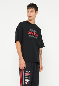 Melnas kokvilnas t-krekls ar īsām piedurknēm, kurā ir baltā un sarkanā krāsā uzraksts "OFFICIAL adidas SUPPLY". Brīva piegriezuma ar apaļu izgriezumu.