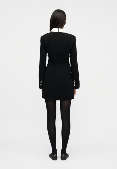 Γυναίκα με μακριά σκούρα μαλλιά, φορώντας εφαρμοστό μαύρο φόρεμα με λευκό γιακά, μαύρες καλσόν και μαύρα φλατ παπούτσια, στέκεται γυρισμένη προς τα πίσω μπροστά σε λευκό φόντο.