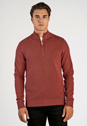 Rode, gestructureerde trui met een hoge kraag en een halve rits. Lange mouwen met ribgebreide boorden en zoom. Draagervaring over een licht hemd en zwarte broek.