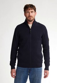Navy zip-up trui met geribbelde structuur, hoge kraag en lange mouwen. Gecombineerd met blauwe spijkerbroek voor een casual look.