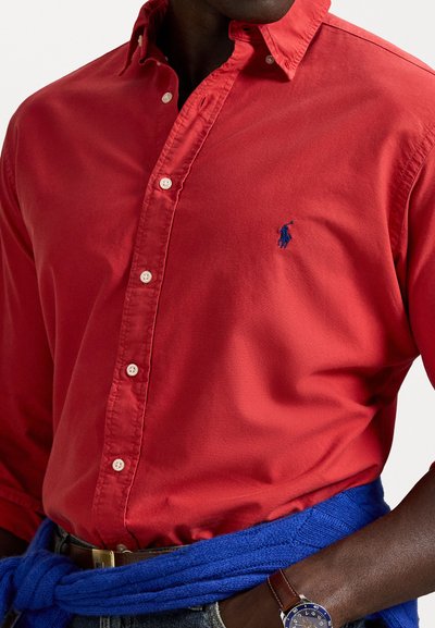 Camisa vermelha de botão, feita de algodão, com um pequeno logotipo azul de polo no peito. Um suéter de malha azul está amarrado à cintura.