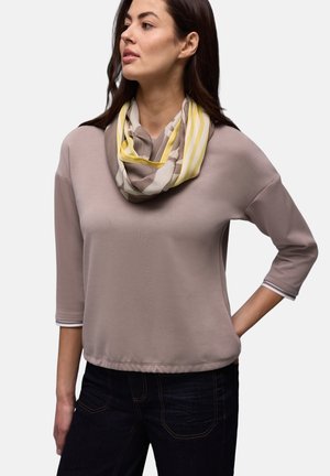 Femme portant un haut mauve à manches trois-quarts et une écharpe jaune à motif beige, avec un jean foncé, posant devant un fond uni.