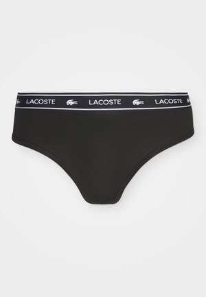 Črne bikini hlačke iz mehkega materiala, s širokim elastičnim pasom, na katerem je v beli barvi napisana oznaka "Lacoste" in krokodilov simbol.