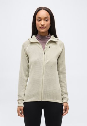 Kobieta w jasnobeżowej dzianinowej kurtce na zamek, nałożonej na fioletowy golf i czarne spodnie, stojąca na jednolitym białym tle.