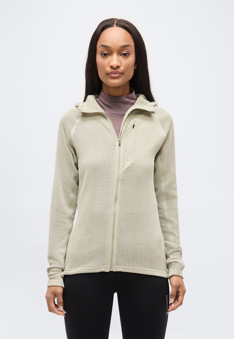 Naine, kes kannab helebeeži lukuga kootud jope üle lilla kõrge kaelusega särgi ja mustade pükstega, seisab ühevärvilise valge tausta ees.