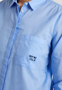 Camicia azzurra chiara con bottoni realizzata in tessuto morbido; presenta una tasca sul petto con scritta ricamata nera "MOP 1967 S.I.H.L.M."