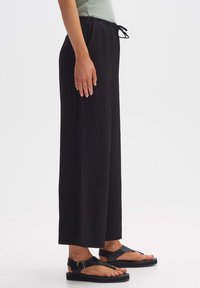 Black pleated wide-leg pants with an elastic waistband and drawstring. Paired with flat black sandals. Simple, modern design.
