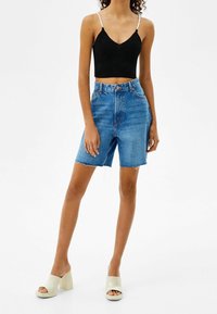 Kvinna som bär en svart kort tanktop med flätade band, högmidjade, knälånga blå jeansshorts och vita klackskor.