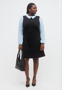 Γυναίκα που φοράει μαύρο αμάνικο φόρεμα πάνω από ανοιχτό μπλε μακρυμάνικο πουκάμισο, κρατώντας μαύρη τσάντα, στέκεται και κοιτάζει πλάγια, μπροστά σε λευκό φόντο.