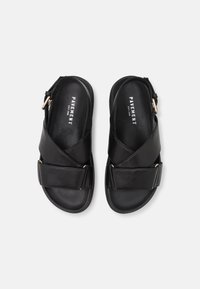 Svart sandaler laget av lær, med kryssremdesign, justerbar spenne, og en glatt overflate med flat såle.