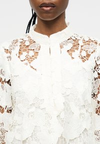 Γυναίκα που φοράει λευκό floral δαντελένιο πουκάμισο με ψηλό γιακά και κουμπωτό μπροστινό μέρος, που δείχνει περίπλοκα σχέδια από δίχτυ και λουλούδια.