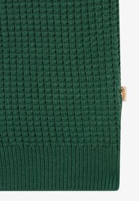 Gros plan sur un tissu tricoté vert foncé avec une texture gaufrée et un ourlet côtelé, comportant une petite étiquette de marque beige sur le côté.