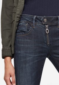 Vaqueros de denim azul oscuro con cierre de cremallera y costuras contrastantes. La tela tiene una textura sutil y un diseño clásico de cinco bolsillos.