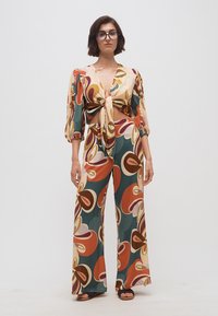 Γυναίκα που φοράει πολύχρωμο τοπ και παντελόνι καμπάνα με αφηρημένο σχέδιο, στέκεται μπροστά σε απλό λευκό φόντο.