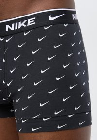 Κοντινό πλάνο ανδρικών εσωρουχων Nike σε μαύρο χρώμα με επαναλαμβανόμενα λευκά λογότυπα Nike swoosh και ζώνη μέσης με το εμπορικό σήμα, σε μοντέλο με σκουρόχρωση επιδερμίδα.