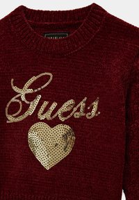 Pull en tricot bordeaux avec le texte "Guess" en sequins dorés et un motif de cœur, doté d'un col côtelé et d'une texture douce.