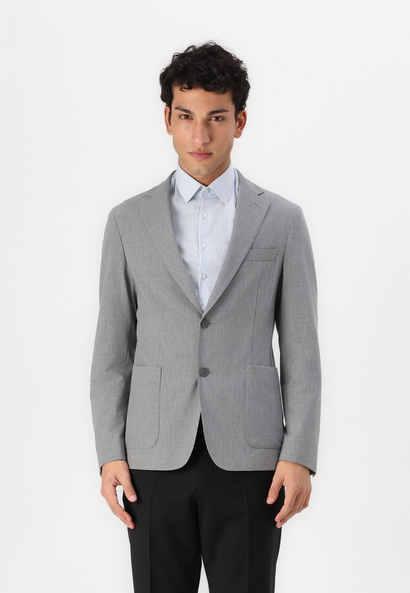 Blazer grigio chiaro con rever a punta, due bottoni e due tasche frontali. Indossato sopra una camicia a righe blu e bianca. Texture liscia.