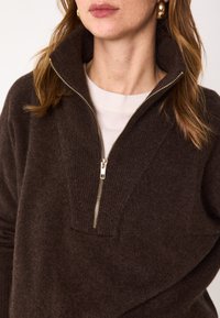Sweater zippé marron en tissu doux avec un col montant et des détails côtelés. Présente une fermeture éclair dorée et une coupe décontractée.