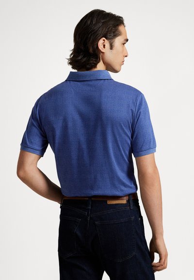 Muž otočený chrbtom, oblečený v modrom textúrovanom polokošeli s krátkym rukávom a tmavých džínsach s hnedým opaskom, na jednoduchom bielom pozadí.