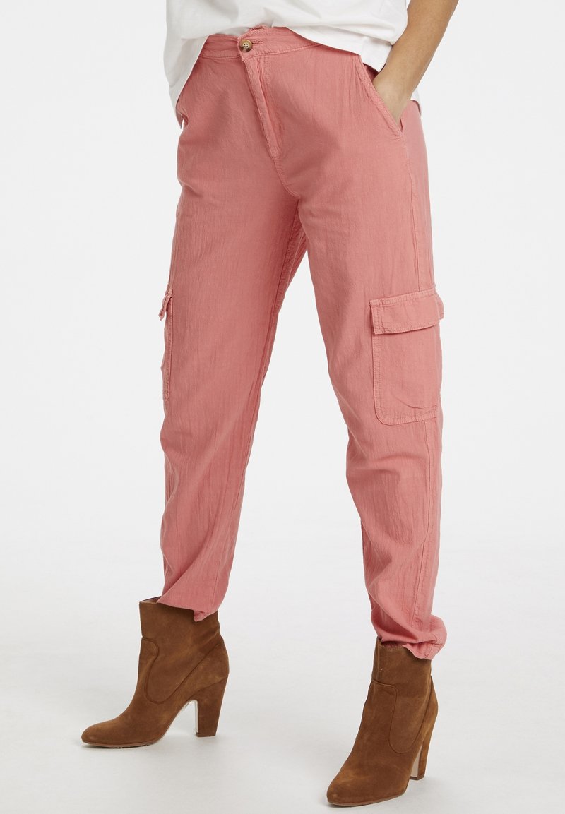 Personne portant un pantalon cargo rose et des bottines à talons en daim marron, debout les mains dans les poches sur un fond blanc.