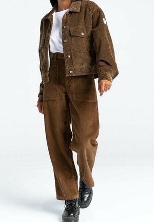 Person iført brun corduroyjakke og bukser med sorte snørestøvler, stående foran en ensfarvet lys baggrund.