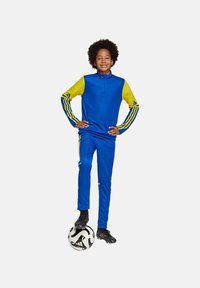 Tuta sportivo blu con accenti gialli, caratterizzato da una parte superiore con zip a quarto e strisce su maniche e pantaloni. Pallone da calcio nero ai piedi.