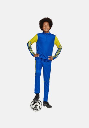 Blauwe sporttracksuit met gele accenten, met een top met kwartrits en strepen op de mouwen en broeken. Zwarte voetbal bij de voeten.
