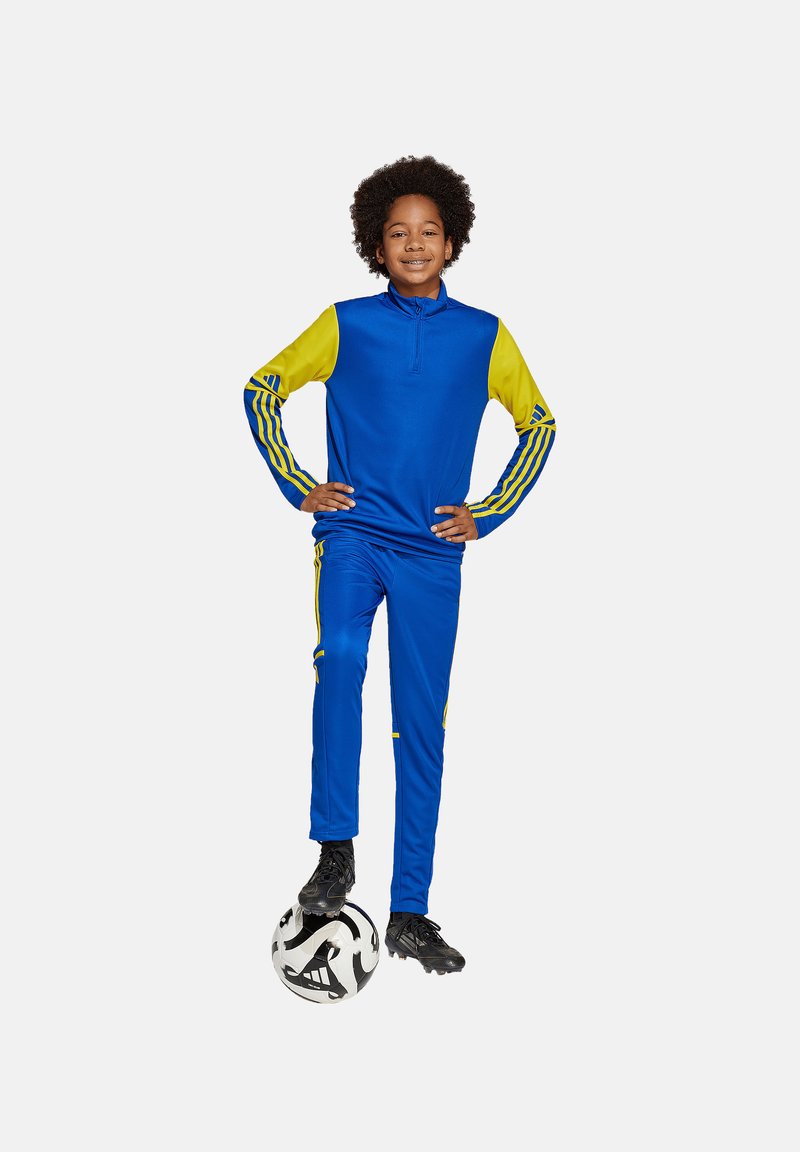Tuta sportivo blu con accenti gialli, caratterizzato da una parte superiore con zip a quarto e strisce su maniche e pantaloni. Pallone da calcio nero ai piedi.