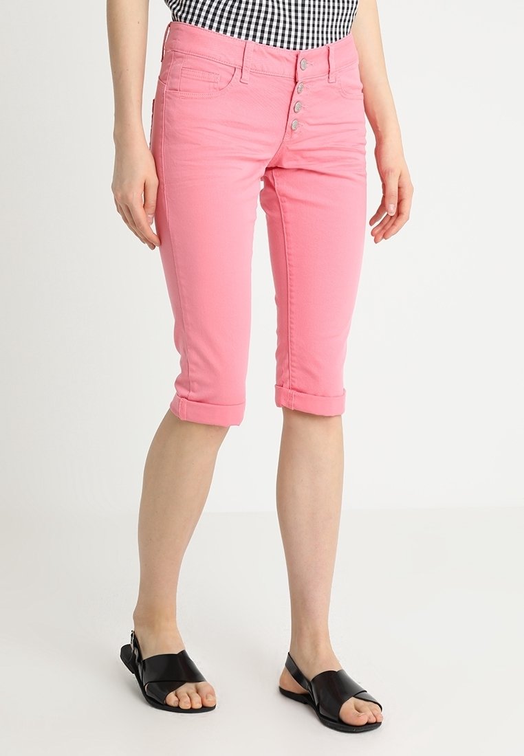 Femme portant un pantalon court rose retroussé avec braguette boutonnée et sandales plates noires ouvertes aux orteils, debout sur fond blanc.