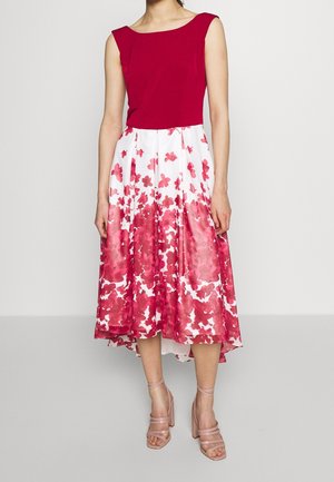 Femme portant une robe sans manches avec un haut rouge et une jupe blanche ornée de motifs floraux rouges, associée à des sandales à talons hauts nude avec des lanières.