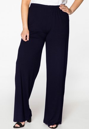 Pantalons larges bleu marine avec une taille élastique, fabriqués en tissu doux. Présentent une texture lisse et une coupe ample, portés avec des sandales à talons noires.