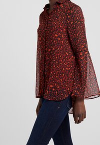 Rød og sort gennemsigtig bluse med leopardmønster, knaplukning foran og klokkeærmer. Letvægtsstof med et flydende design.