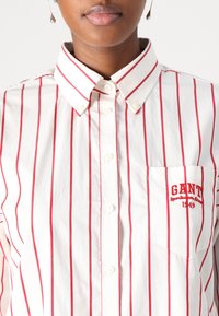 Camicia a maniche lunghe a righe con strisce rosse su sfondo bianco. Include una tasca sul petto con logo ricamato in rosso. Materiale in cotone.