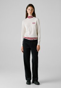 Maglione bianco lavorato a maglia con righe rosse e navy sul colletto e sull'orlo, caratterizzato dal logo "Red Bull Racing". Abbinato a pantaloni in corduroy neri.