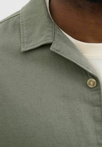 Camisa verde oliva con cuello, hecha de tela texturizada, que presenta un botón único y detalles de costura visibles. Superpuesta sobre una blusa color crema.