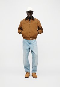 Άντρας που φοράει μπεζ μπουφάν bomber, ανοιχτό μπλε τζιν, μπεζ μπότες και λευκό καπέλο, στέκεται με τα χέρια στις τσέπες σε λευκό φόντο.