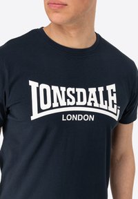 Mornarsko plava pamučna majica s velikim bijelim logotipom "LONSDALE LONDON", okruglim izrezom i kratkim rukavima. Glatka tekstura i ležeran kroj.