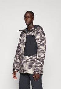 Chaqueta de hombre con un patrón en espiral en negro y gris, cierre de cremallera y un panel en negro contrastante en el pecho y el brazo. Presenta una capucha.
