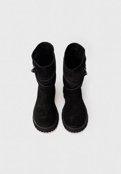 Ζευγάρι μαύρες σουέντ ankle boots με χοντρές ελαστικές σόλες και λουράκια με αγκράφα στο πλάι, προφίλ από πάνω σε λευκό φόντο.