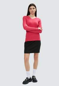 Maglione rosa a coste con maniche lunghe, abbinato a una gonna nera e mocassini neri con accentuazione in catena dorata. Calzini bianchi visibili.