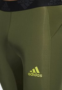 Olive zielone legginsy sportowe z czarnym elastycznym pasem, na którym znajduje się subtelne logo Adidas w jasnej zieleni. Wykonane z gładkiego, elastycznego materiału.