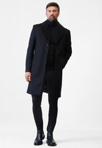 Schwarzer Wollmantel mit einem dunklen Pelzkragen, in taillierter Form, mit fünf Knöpfen und seitlichen Taschen. Getragen über einem schwarzen Rollkragenpullover.