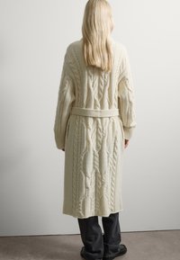 Largo cárdigan de punto cable en color crema con cintura ceñida, que presenta patrones intrincados y mangas anchas, combinado con pantalones oscuros.