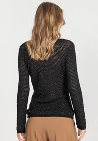 Kobieta o falujących blond włosach ma na sobie czarny, wzorzysty sweter z długim rękawem oraz jasnobrązowe spodnie, stoi tyłem na jednolitym tle.