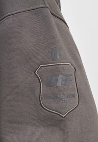 Sivi materijal dukserice sadrži zakrpu u obliku štita s utisnutim tekstom "DEF" i "WORKATION COLLECTION FW23", uz dodatak prošivenog obruba.