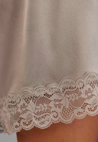 Satin nattkläder med en nederkant av intrikat blommig spets i ljusbeige, som visar en slät textur och en delikat scalloped kantdetalj.