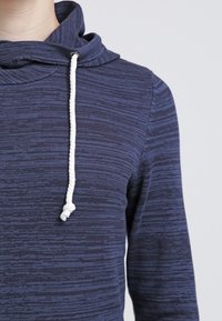 Sweat à capuche bleu marine en tissu texturé, doté d'un cordon de serrage avec des accents blancs et d'une coupe décontractée. Design minimaliste sans motifs.